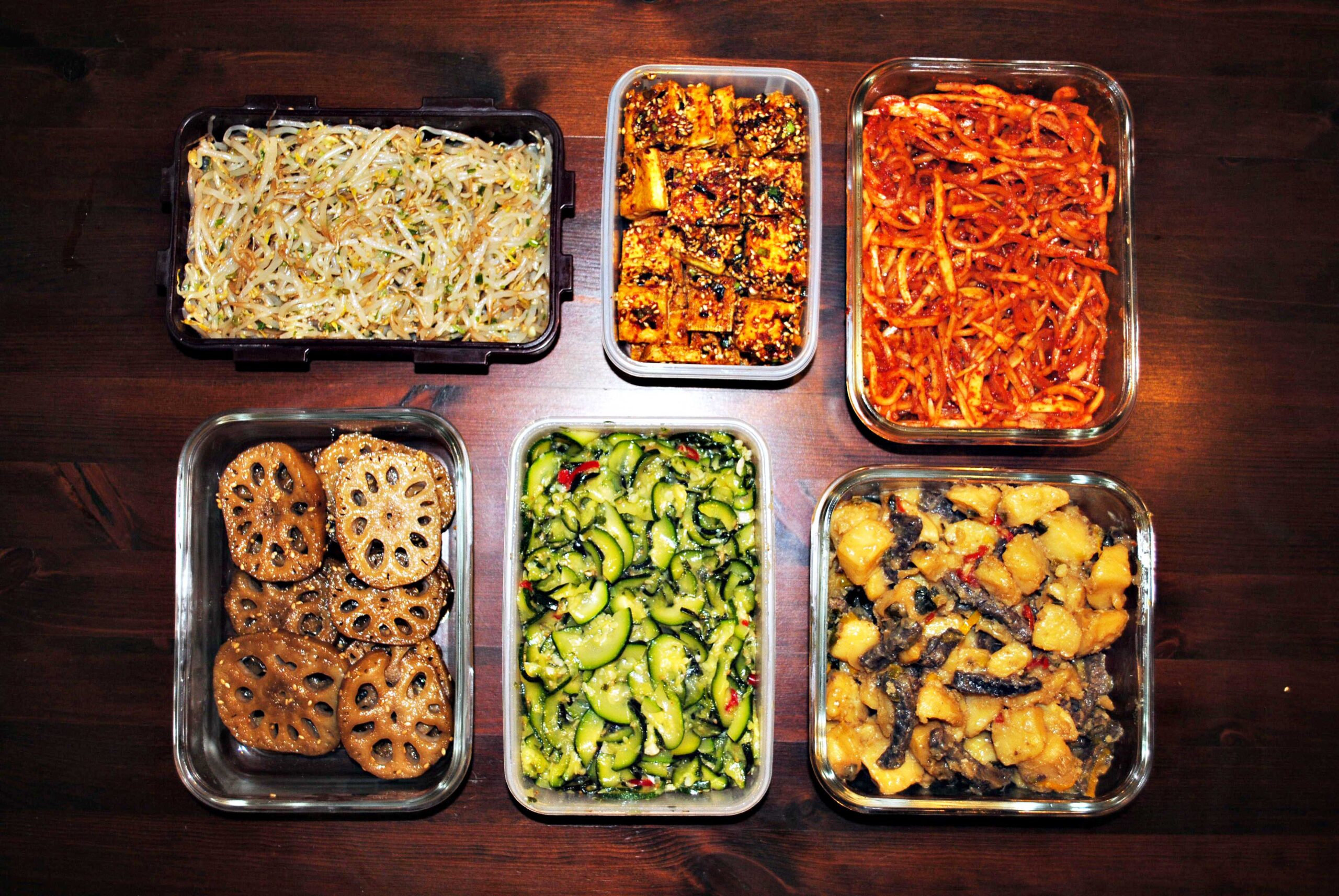 Sechs koreanische Banchan in Boxen: Sprossen-Muchim, Tofu-Buchim, Rettich-Muchim, Lotus-Jorim, Zucchini-Namul und Kartoffel-Rindfleisch-Jorim
